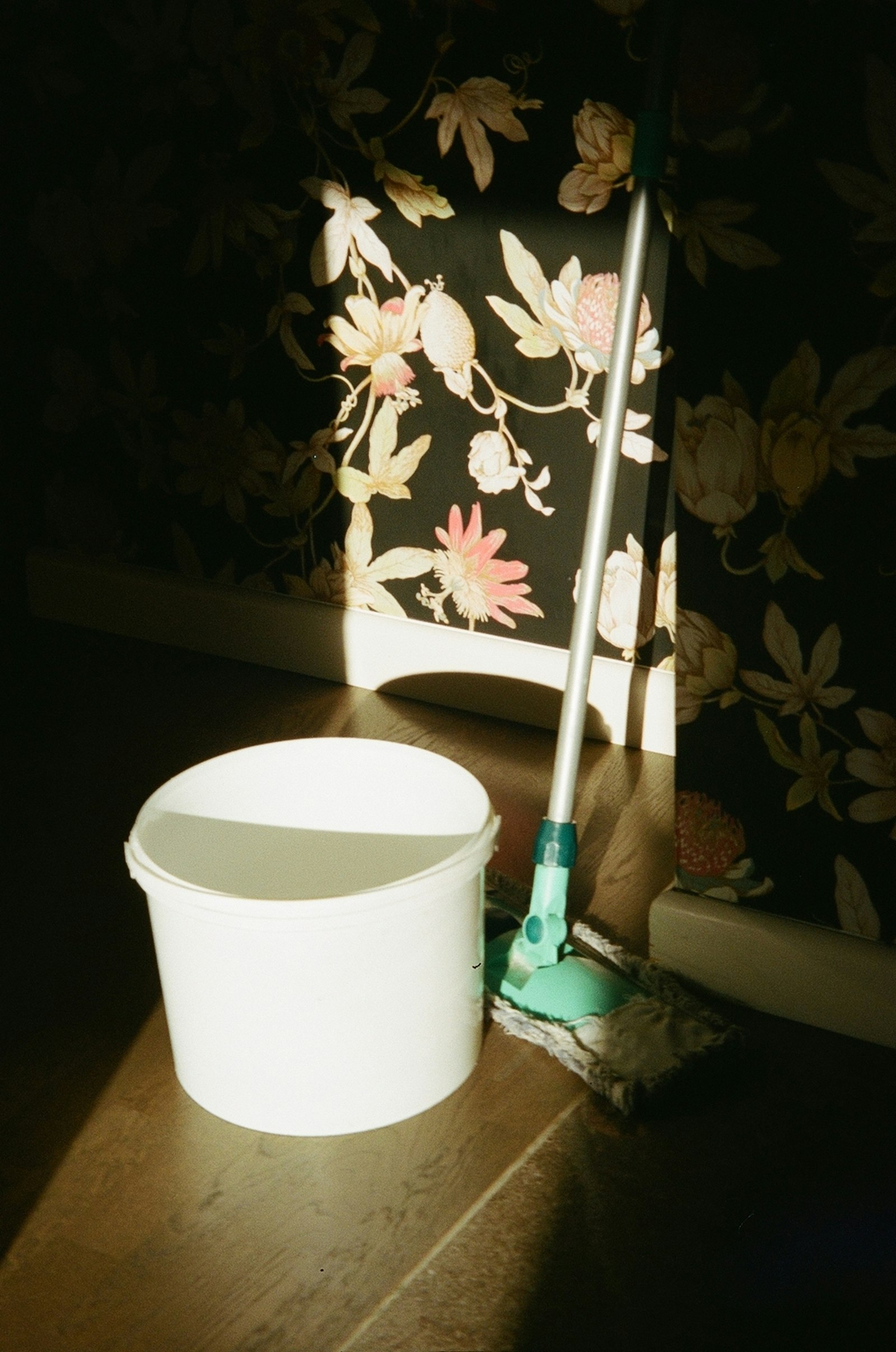 A white bucket and a mop standing in a patch of sunlight against a patterned wall.